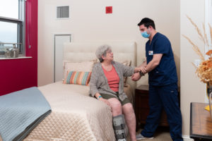 A healthcare worker in scrubs assists an elderly woman sitting on a bed in a well-lit room, both smiling and holding hands.