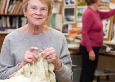 Elderly woman with a joyful expression knitting a pale yellow garment, sitting in a workshop with another woman painting in the background.