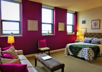 A stylish bedroom with bright fuchsia walls, featuring two beds with patterned covers, framed artwork, and multiple windows.