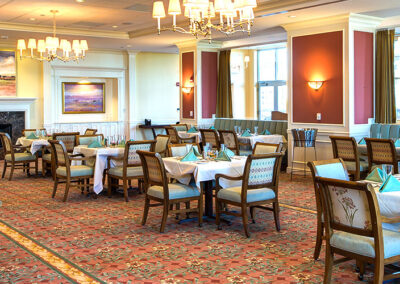 Elegant dining room with set tables, patterned carpet, and a fireplace, bathed in natural light from large windows.