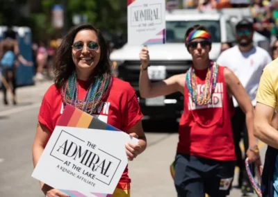 A woman smiling and holding a sign reading "the admiral at the lake" at a sunny pride parade, accompanied by another participant in the background.
