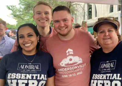 Four cheerful adults posing closely for a photo, two wearing navy-themed t-shirts labeled "the admiral," and one in a pale pink "andersonville midsommarfest" shirt.