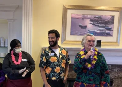 Three people, one wearing a lei, stand together in a room with a boat painting in the background. two are in floral shirts and one in a black outfit.