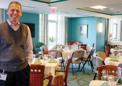 Man smiling in a restaurant with tables set for dining, teal walls, and large windows.