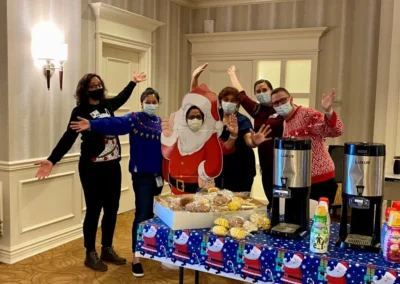 A group of six people celebrating at a holiday party, dressed in festive attire and posing joyfully around a table with snacks and drinks.