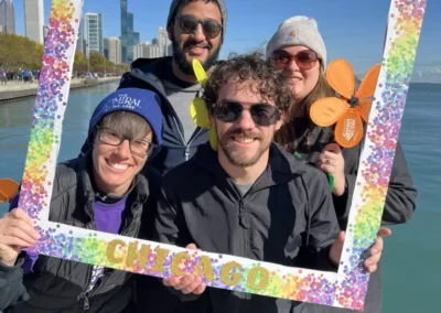 Four adults smiling while holding a colorful "chicago" themed photo frame by a lakeside with a city skyline in the background.