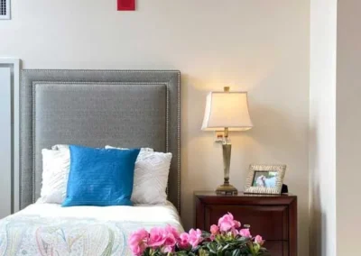 Elegant bedroom with a grey tufted headboard, a bright blue pillow among white ones, a wooden bedside table with a lamp, and pink flowers in the foreground.