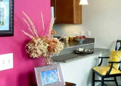 A vibrant room corner featuring a magenta wall, a granite countertop with a flower arrangement and kitchen appliances, and a yellow chair nearby.