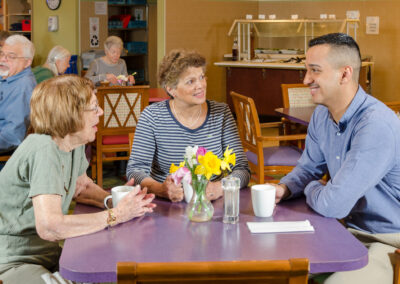 Three elderly individuals and a young man conversing around a table in a care home, with others in the background.