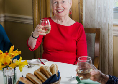 Elderly woman in a red blouse smiling, holding a glass of wine at a dining table with flowers and bread in focus.