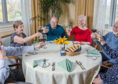 Group of five elderly people toasting with wine at a round dining table, smiling in a brightly lit room with a scenic window view.