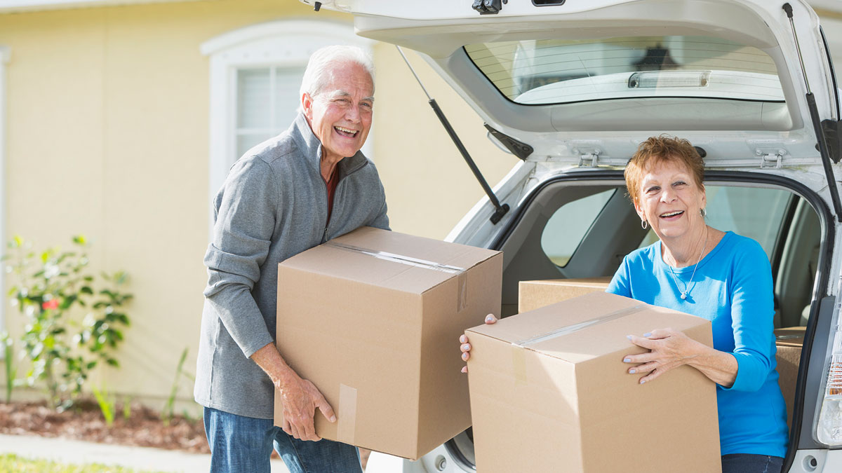 Elderly couple smiling and lifting cardboard boxes from the trunk of a car in front of a house.